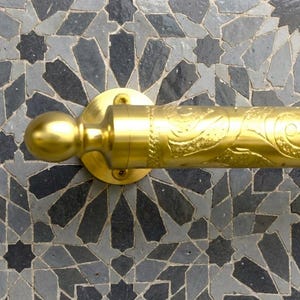 Może przedstawiać: Złoty metalowy wieszak na ręczniki z dekoracyjnym kwiatowym wzorem. Drążek jest zamontowany na okrągłej płycie za pomocą śrub, na tle wzoru mozaiki w odcieniach szarości i czerni.