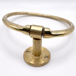 Op de afbeelding: Een messing handdoekring met een cirkelvormig ontwerp. De ring is bevestigd aan een ronde basis met een korte, cilindrische paal. De ring heeft ge&euml;tste bloempatronen aan de uiteinden. De armatuur is gemaakt van messing en heeft een glanzende afwerking.