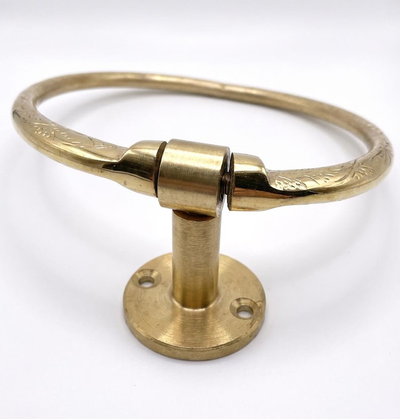 Op de afbeelding: Een messing handdoekring met een cirkelvormig ontwerp. De ring is bevestigd aan een ronde basis met een korte, cilindrische paal. De ring heeft ge&euml;tste bloempatronen aan de uiteinden. De armatuur is gemaakt van messing en heeft een glanzende afwerking.