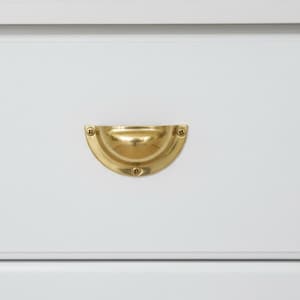 Puede incluir: Primer plano de un cajón blanco con un tirador de cajón de latón pulido en forma de media luna. El tirador está asegurado con tornillos pequeños y tiene una superficie reflectante. El herraje añade un toque de elegancia al cajón blanco.