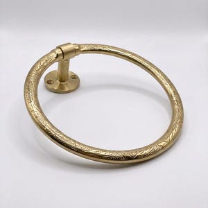 Op de afbeelding: Een goudkleurige handdoekring met een sierlijk bloemmotief. De cirkelvormige ring is bevestigd aan een ronde basis met een korte, cilindrische arm. Het ontwerp is vintage en geschikt voor een badkamer.