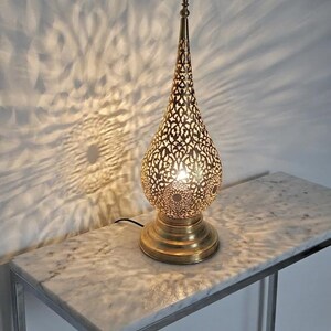 May include: A decorative gold-coloured table lamp with a detailed, teardrop-shaped shade. The lamp's intricate cut-out design casts a patterned light on the wall. It is placed on a marble-topped table. The lamp's height is approximately 40 cm.