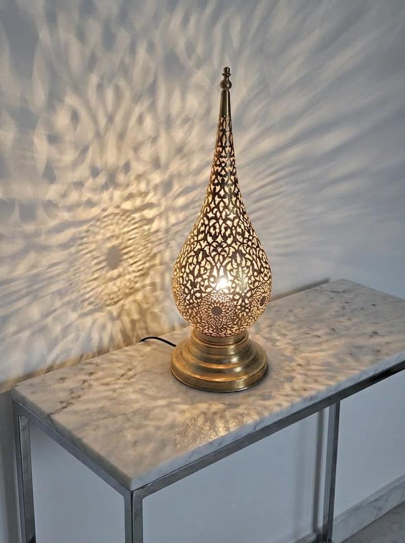 May include: A decorative gold-coloured table lamp with a detailed, teardrop-shaped shade. The lamp's intricate cut-out design casts a patterned light on the wall. It is placed on a marble-topped table. The lamp's height is approximately 40 cm.