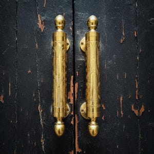 May include: A pair of gold-coloured door handles with detailed patterns, mounted on a distressed black wooden door. The handles have a cylindrical shape with decorative finials at the top and bottom.
