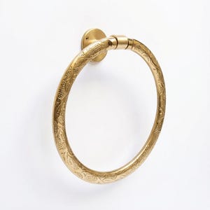 Op de afbeelding: Een messing handdoekring met een sierlijk bloemmotief. De ring is gemonteerd op een cirkelvormige basis en is ontworpen om een handdoek vast te houden. Het messing heeft een warme, gouden tint en het ontwerp is in het metaal ge&euml;tst.