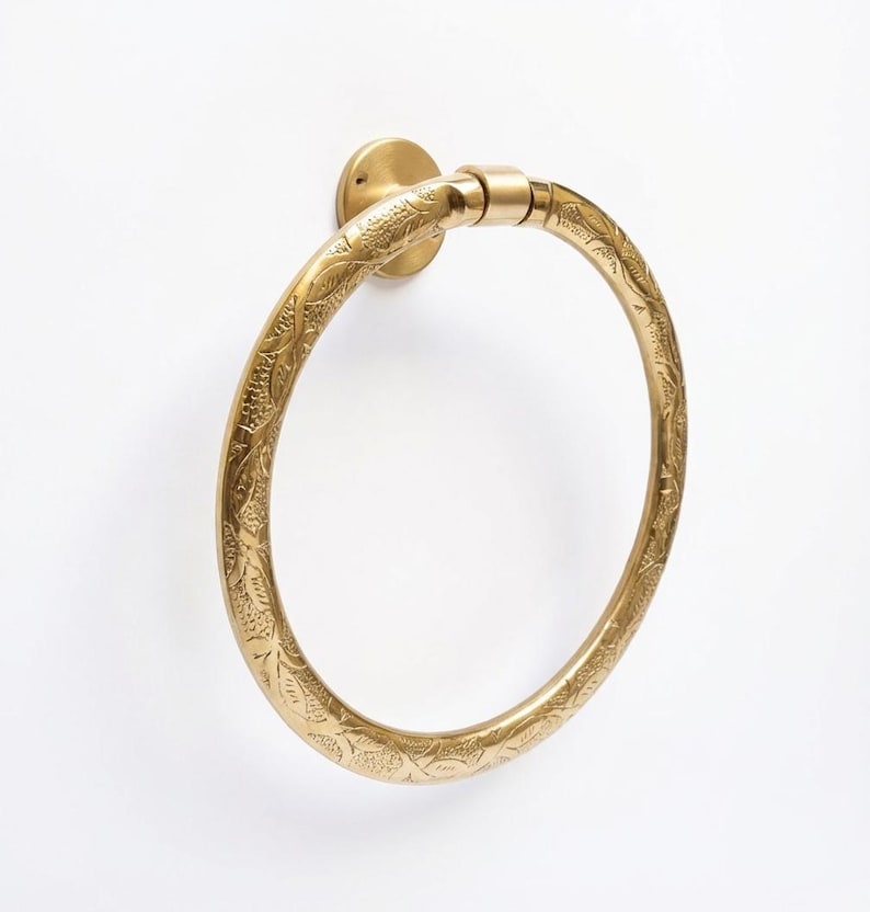 Op de afbeelding: Een messing handdoekring met een sierlijk bloemmotief. De ring is gemonteerd op een cirkelvormige basis en is ontworpen om een handdoek vast te houden. Het messing heeft een warme, gouden tint en het ontwerp is in het metaal ge&euml;tst.