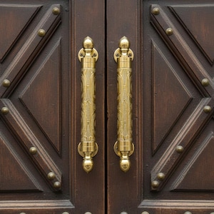 Puede incluir: Primer plano de una puerta de madera marrón oscuro con dos manijas doradas ornamentadas. Los paneles de la puerta presentan un diseño geométrico con pequeños tachuelas redondas de color latón. Las manijas tienen patrones florales detallados.