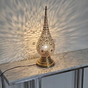 May include: A decorative gold-coloured table lamp with a pierced, teardrop-shaped shade. The lamp sits on a round base and casts intricate light patterns on the wall. The lamp is plugged into an electrical outlet.