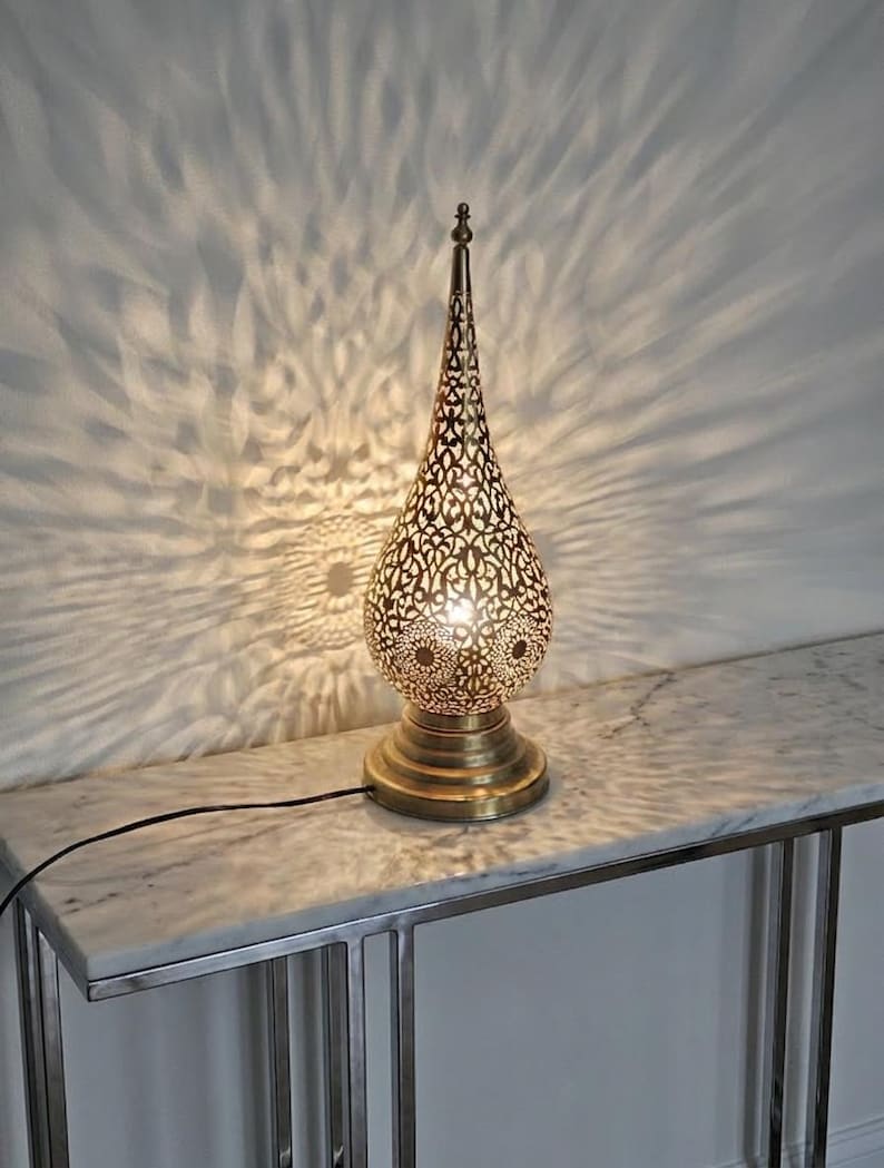 May include: A decorative gold-coloured table lamp with a pierced, teardrop-shaped shade. The lamp sits on a round base and casts intricate light patterns on the wall. The lamp is plugged into an electrical outlet.