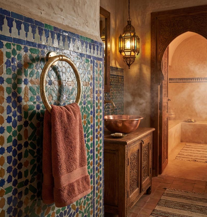 Op de afbeelding: Badkamer met een gouden handdoekring met een koraalkleurige handdoek. De muur is versierd met kleurrijke moza&iuml;ektegels. Een houten kast met een koperen wastafel en een hangende lantaarn maken de sc&egrave;ne compleet.