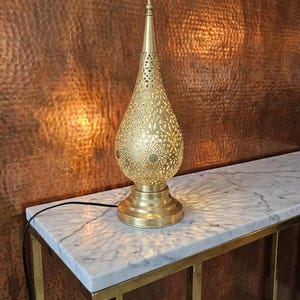 May include: A decorative gold table lamp with a pierced, teardrop-shaped shade. The lamp is on a marble-topped table with a gold frame, set against a textured, copper-toned wall. The lamp's design casts patterned light.