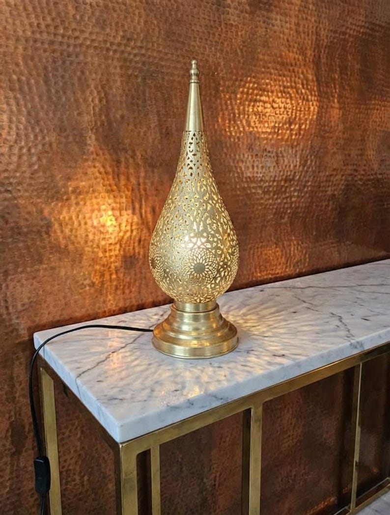 May include: A decorative gold table lamp with a pierced, teardrop-shaped shade. The lamp is on a marble-topped table with a gold frame, set against a textured, copper-toned wall. The lamp's design casts patterned light.
