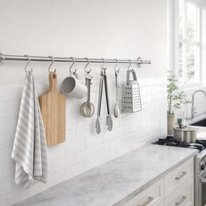 May include: A stainless steel kitchen utensil rack with various tools hanging from it. Items include a wooden cutting board, a mug, tongs, a grater, and a striped towel. The kitchen has white cabinets and a marble countertop.