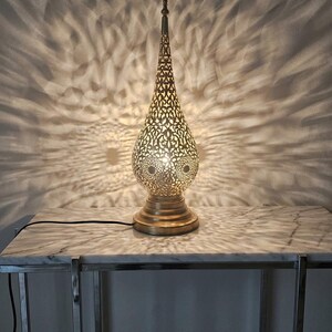 May include: A decorative gold-toned table lamp with a detailed, cut-out design. The lamp, with its teardrop shape, is placed on a marble-effect table, creating a patterned light effect on the wall. The lamp has a round base and a pointed top.