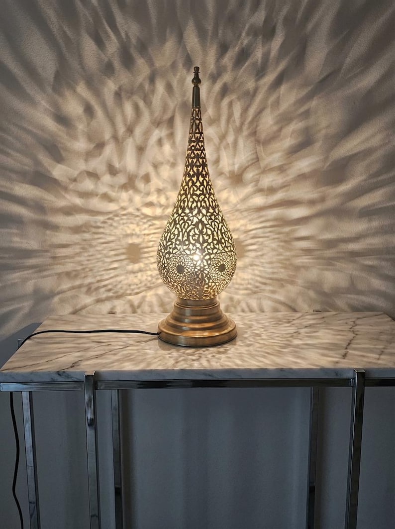 May include: A decorative gold-toned table lamp with a detailed, cut-out design. The lamp, with its teardrop shape, is placed on a marble-effect table, creating a patterned light effect on the wall. The lamp has a round base and a pointed top.