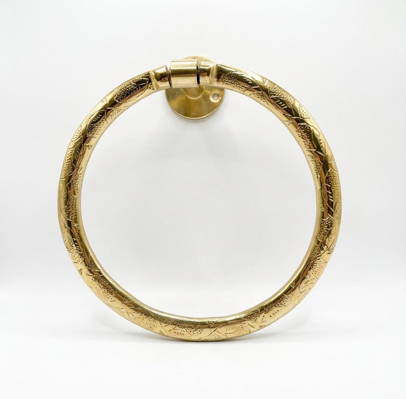 Op de afbeelding: Een messing handdoekring met een ge&euml;tst bloemmotief. De cirkelvormige ring is bevestigd aan een ronde wandbevestiging. Het metaal heeft een warme, gouden tint en het ontwerp is symmetrisch.