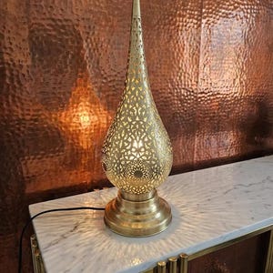 May include: A gold-coloured, ornate table lamp with a teardrop-shaped shade featuring intricate cut-out patterns. The lamp sits on a round base and emits a warm glow. The lamp is approximately 61 cm tall.