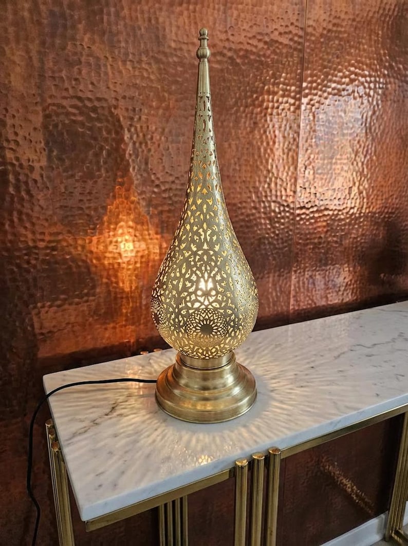 May include: A gold-coloured, ornate table lamp with a teardrop-shaped shade featuring intricate cut-out patterns. The lamp sits on a round base and emits a warm glow. The lamp is approximately 61 cm tall.