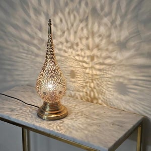 May include: A decorative gold-coloured table lamp with a pierced metal shade, creating a patterned light effect on the wall. The lamp has a teardrop shape and sits on a round base. The lamp is on a marble-topped table with a gold metal frame.