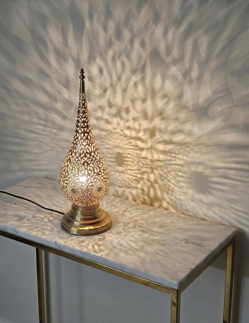 May include: A decorative gold-coloured table lamp with a pierced metal shade, creating a patterned light effect on the wall. The lamp has a teardrop shape and sits on a round base. The lamp is on a marble-topped table with a gold metal frame.