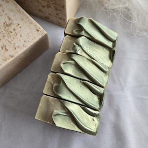 May include: Handmade soap bars in green and beige tones. The top of the soap features a decorative, swirled pattern in a metallic green. Several bars are stacked, with more bars visible in the background, creating a natural aesthetic.