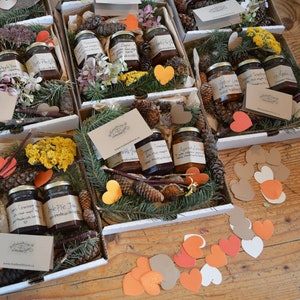 May include: Assortment of small glass jars filled with jam, arranged in white boxes with brown paper hearts and pine branches. The jars have handwritten labels with names like "Cranberry Jam" and "Apple Pie Jam".