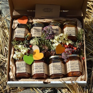 May include: A presentation box containing jars of preserves, decorated with flowers and heart-shaped cutouts. The jars are labelled with handwritten names such as "Cranberry" and "Green Gage Plum". The box is presented on a bed of straw.