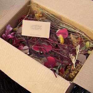 Op de afbeelding: Een kartonnen doos gevuld met gedroogde bloemen, takjes en rode papieren hartjes. De doos bevat een kaart met de tekst "Made with Love & Deliciously Yours" met een websiteadres.