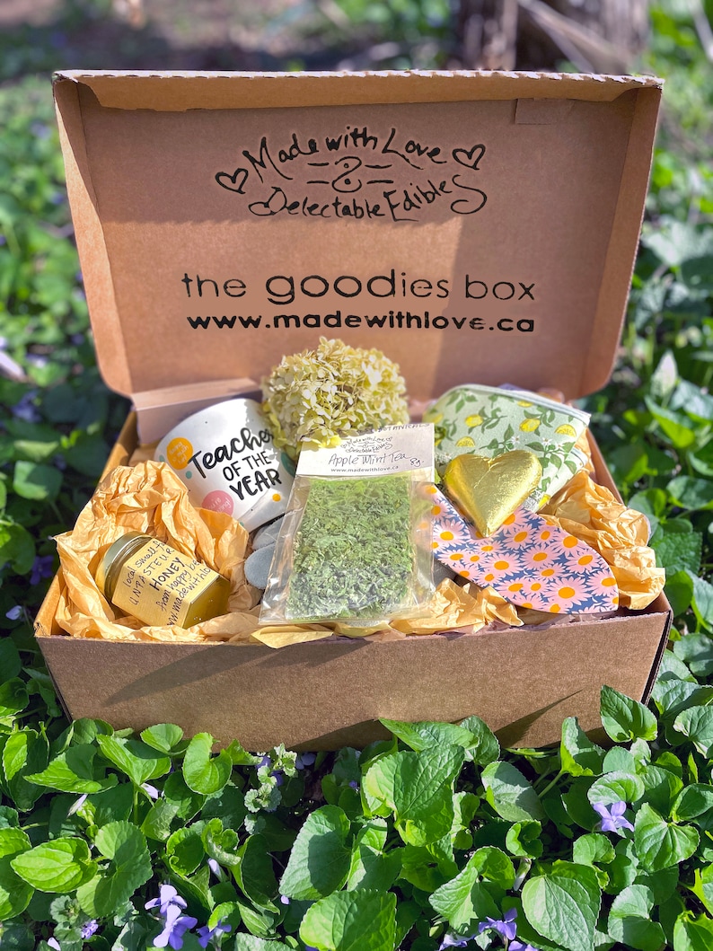 Puede incluir: Una caja de regalo marr&oacute;n abierta, llena de varios art&iacute;culos, incluyendo una taza con la inscripci&oacute;n "Teacher of the Year", un tarro de miel, una bolsita de t&eacute; de menta y una bolsa con estampado floral. La caja est&aacute; etiquetada "Made with Love".