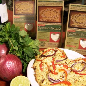 May include: Two packages of rosemary focaccia mix from Made with Love & Relectable Edibles. The packages are brown with a heart-shaped logo and the text "Made with Love & Relectable Edibles" and "www.madewithlove.ca". The packages are on a wooden table with a plate of focaccia bread topped with red peppers, red onions, and white cheese.