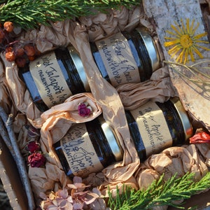 May include: A rustic gift box containing four jars of jam with gold lids and handwritten labels. The jars are packed in brown paper with dried flowers, foliage, and birch bark. The labels read "Spiced Jalapeño", "Pear & Rowan", "Pepper Apple", and "Fairy Fruit & Berries".