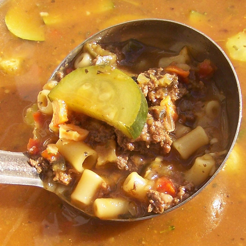 Op de afbeelding: Een close-up van een soeplepel gevuld met een stevige soep. De soep bevat pasta, gehakt, groenten en een schijf komkommer. De soep zit in een grote pan en de soeplepel wordt uit de pan gelicht.