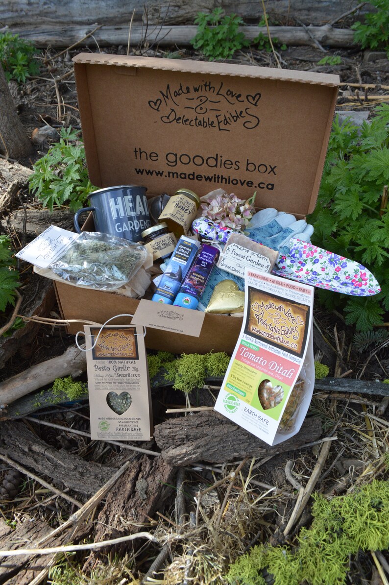 Puede incluir: Una caja de cart&oacute;n marr&oacute;n, "the goodies box", llena de varios art&iacute;culos de jardiner&iacute;a y alimentos. Incluye una taza, tarros de miel, paquetes de especias y guantes de jardiner&iacute;a. La caja est&aacute; rodeada de follaje natural.