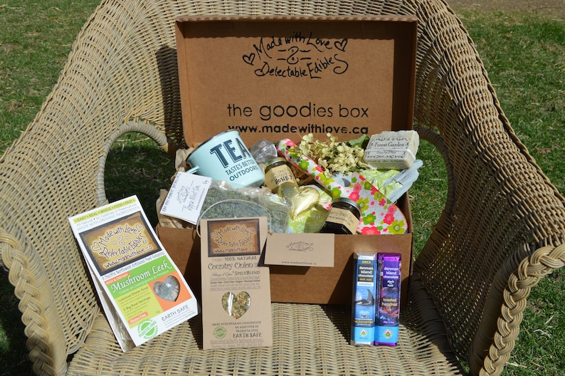 Puede incluir: Una silla de mimbre sostiene una caja de cart&oacute;n marr&oacute;n etiquetada como "the goodies box" llena de varios art&iacute;culos. En el interior hay una taza de color turquesa, paquetes de hierbas, jabones y chocolates. La caja est&aacute; decorada con el texto "Made with Love & Delectable Edibles".