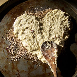 May include: A heart-shaped dough spread on a baking sheet with sesame seeds sprinkled around it. A wooden spoon is resting on the dough.