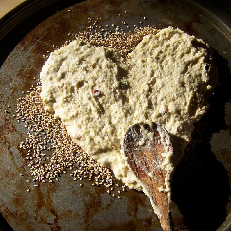 May include: A heart-shaped dough spread on a baking sheet with sesame seeds sprinkled around it. A wooden spoon is resting on the dough.