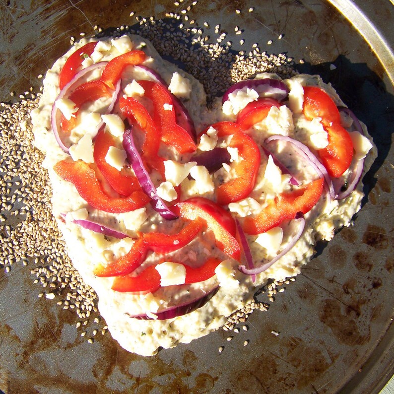 May include: A heart-shaped pizza crust topped with red bell peppers, red onion, and white cheese. The pizza is on a baking sheet sprinkled with sesame seeds.