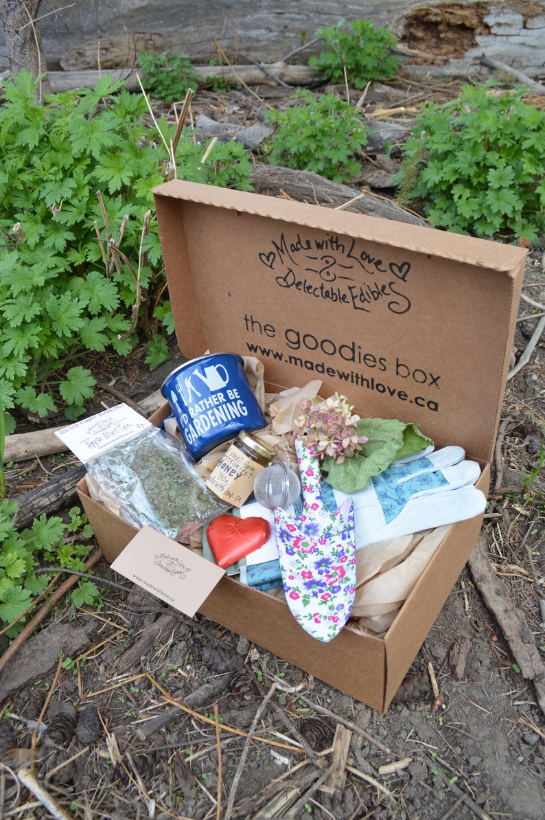 Puede incluir: Una caja de cart&oacute;n marr&oacute;n llena de art&iacute;culos de jardiner&iacute;a. Incluye una taza azul con "I'd Rather Be Gardening", una paleta floral, guantes, miel, t&eacute; y un objeto en forma de coraz&oacute;n. La caja est&aacute; etiquetada como "the goodies box".