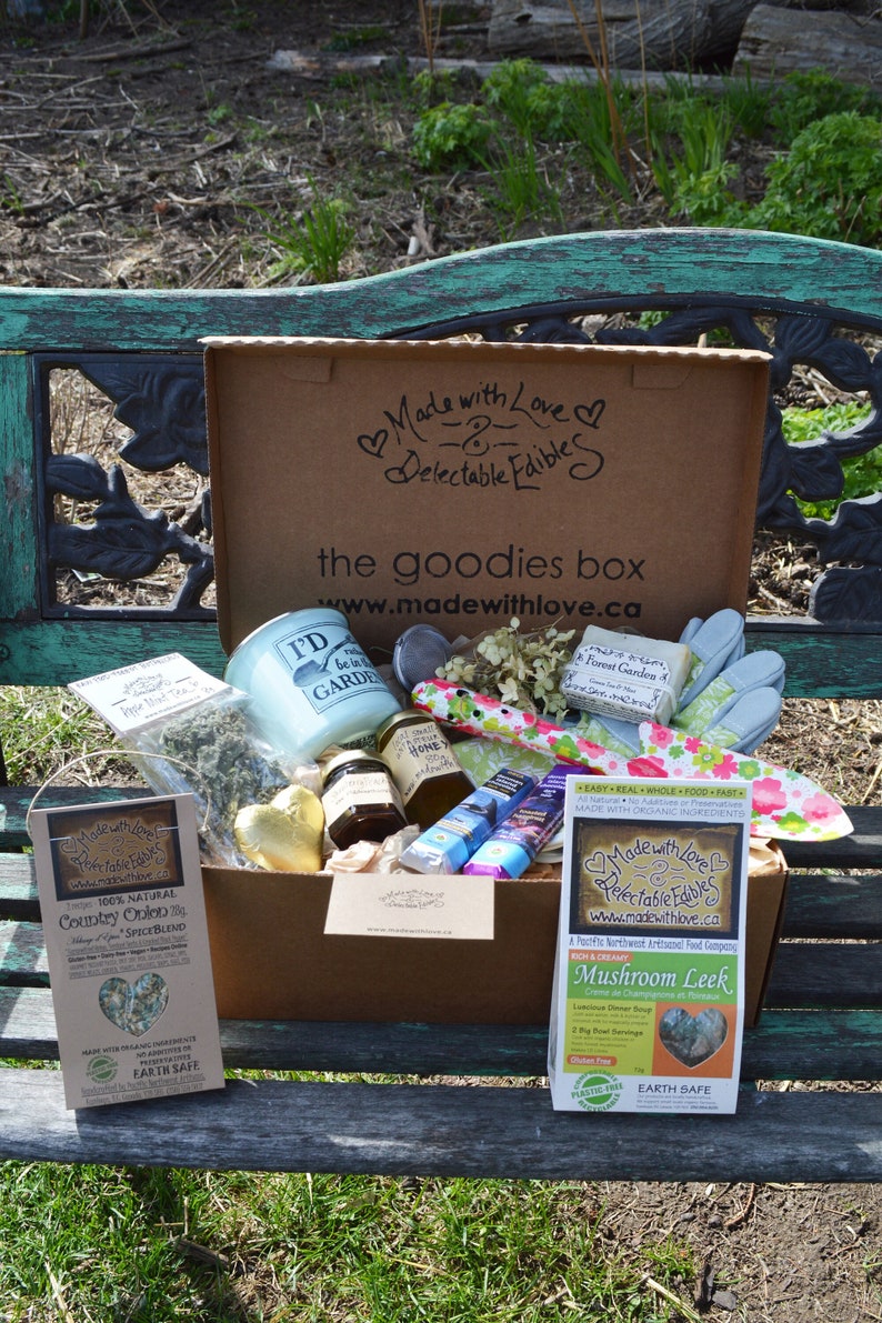 Puede incluir: Una caja de regalo de cart&oacute;n, con la marca "Made with Love & Delectable Edibles", llena de varios productos alimenticios, incluyendo miel y una vela. La caja est&aacute; sobre un banco verde, al aire libre.