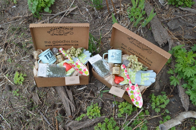 Puede incluir: Dos cajas de regalo abiertas con art&iacute;culos de jardiner&iacute;a. Cada caja contiene una taza, guantes, una paleta y otros objetos peque&ntilde;os. Las cajas est&aacute;n sobre papel marr&oacute;n, con el texto "the goodies box" y "Made with Love" en la caja.
