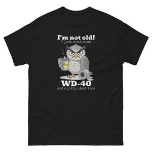 以下が含まれることがあります： WD-40の缶とダクトテープのロールを持ったフクロウの漫画が描かれた黒いTシャツ。テキストには「I'm not old! I just need some WD-40 and a little duct tape」と書かれています。フクロウは不機嫌そうな表情で、灰色の羽と黄色い足が特徴です。