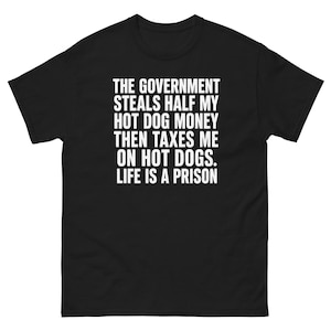 May include: A black short-sleeved t-shirt featuring white text that reads: THE GOVERNMENT STEALS HALF MY HOT DOG MONEY THEN TAXES ME ON HOT DOGS. LIFE IS A PRISON. The shirt has a crew neckline and a standard fit, displaying the text centered on the chest.