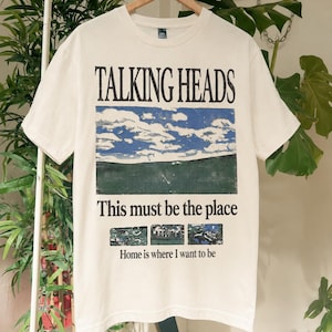 Puede incluir: Una camiseta de manga corta color blanco roto con un estampado gráfico que incluye el texto TALKING HEADS, This must be the place y Home is where I want to be. El diseño presenta una ilustración de un paisaje con cielo azul y nubes.