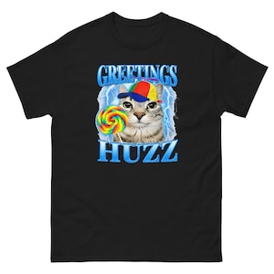 May include: A black t-shirt featuring a graphic of a cat wearing a multi-colored propeller cap and holding a rainbow-colored lollipop. The design includes blue lightning bolts in the background and the text GREETINGS HUZZ in blue lettering.