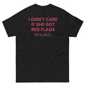 Może przedstawiać: Czarny t-shirt z krótkim rękawem z różowym napisem na plecach: I DON'T CARE IF SHE GOT RED FLAGS I'M A BULL. Koszulka ma okrągły dekolt i standardowy krój, wyraźnie prezentując tylną część odzieży.