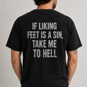 Può includere: Una t-shirt nera a maniche corte con il testo IF LIKING FEET IS A SIN, TAKE ME TO HELL stampato sul retro in un carattere sans-serif grigio chiaro dall'aspetto usurato. Il capo presenta un girocollo e una vestibilità standard rilassata.