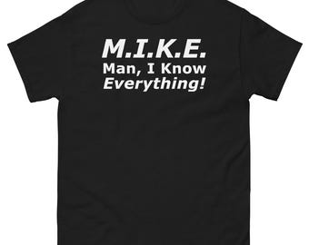Op de afbeelding: Zwart T-shirt met de witte tekst "M.I.K.E. Man, I Know Everything!" op de borst. Het shirt heeft een ronde hals.