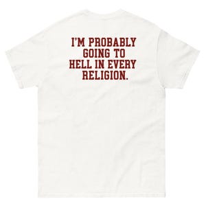 Puede incluir: Una camiseta blanca de manga corta con letras mayúsculas de color rojo oscuro en la parte posterior que dice: I'M PROBABLY GOING TO HELL IN EVERY RELIGION. La prenda tiene cuello redondo y un corte estándar, mostrando el texto claramente.