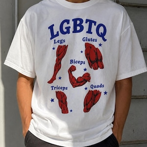 Puede incluir: Una camiseta blanca de manga corta con el texto azul LGBTQ en la parte superior. Debajo hay ilustraciones anatómicas rojas de músculos etiquetadas como Legs, Glutes, Biceps, Triceps y Quads en azul, con pequeñas estrellas azules dispersas.