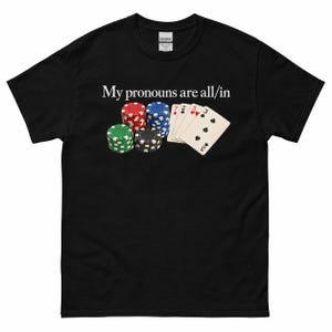 May include: A black short-sleeve t-shirt featuring a graphic of four stacks of colorful poker chips and a hand of playing cards. Above the graphic, the white text reads, My pronouns are all/in. The shirt has a standard crew neckline and short sleeves.